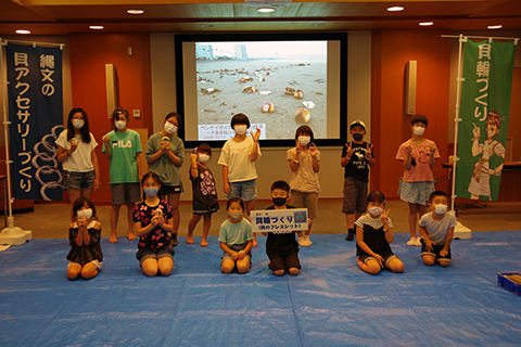 緑の貝輪づくりとと書かれたのぼりがあり、子どもたちが二列に並び前列の一人の男の子が貝輪づくりというプレートを持っている集合写真