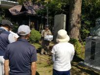 銅像や石碑が建っている前に立つ男性が話をしているのを聞いている参加者たちの後ろ姿の写真