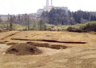 奥に工場のようなものが見え、四角く溝が掘られた手前に彫った土を盛った山がある写真