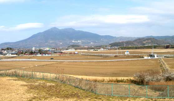 一面が茶色い土畑で手前には緑色のフェンスがあり、遠くに山を望むのどかな風景の写真