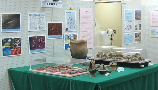 展示室の様子、白い壁には8枚のパネルがあり緑の布が敷かれた台の上には土器とケース入りの石のようなものが並ぶ写真