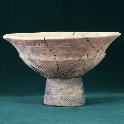 深い緑の背景で、茶碗型の鉢の下に円柱状の台がついている継ぎ目のある薄茶色の台付浅鉢形土器の写真