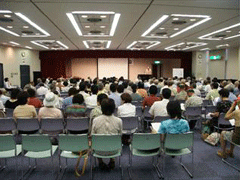 セミナー会場のような場所で1人が前に登壇し、たくさんの人がいすに座って聞いている様子の写真