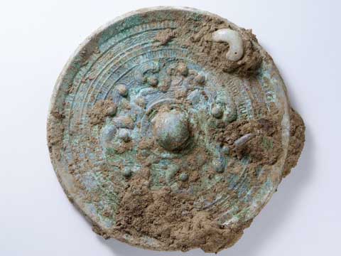 円形で色は青緑色、全体に茶色い土のようなものが付着しており、右上に勾玉のようなものが装飾されている和鏡の写真