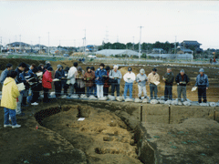 遺跡を囲って見学をしている20人近くの人が横1列に並んでいる写真