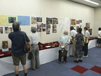 室内の壁に写真や説明などが貼られていて、その下の棚にも展示物が並べられ、それらを眺めている人が7名ほど写っている写真