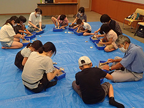 ブルーのシートに座り複数の親子が貝を割り、穴を広げる作業をしている写真