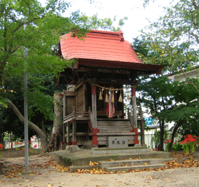 赤い屋根の祭神の周辺にヒガンバナや草木が咲き、落ち葉が落ちている写真