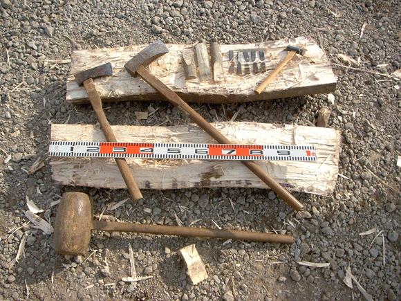 縦に半分に裂けた丸太が割れ目を上に横たわり、その上に小さな斧や木製、鉄製の様々なクサビ、メジャーなどの道具が置かれ、側には木製の大きなトンカチも置かれている様子の写真