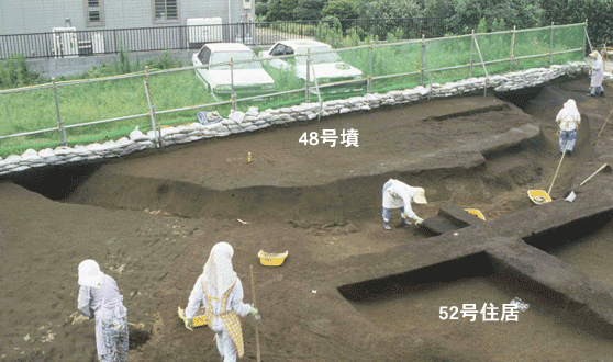 古墳と竪穴住居跡の周溝で数名の作業員が発掘調査をしている写真
