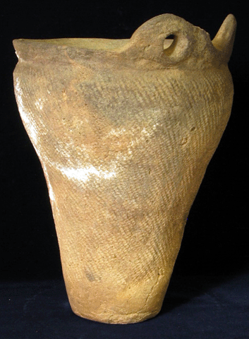 薄茶色で上が広い花瓶のような形で表面に斜めの線の模様が一面に入っており、右上部に穴の空いた持ち手の様なものがついている土器の写真