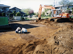 土の上に2人がしゃがみ込みその脇でショベルカーが土を掘っている写真