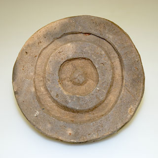 丸くて焦げ茶色の円盤の中に溝のような線が三重になっている土器の写真