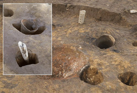 一面土の中央部が一段低くなっており、低くなっているところに穴が点在し、いくつかの穴には石の棒のようなものや筒状の何かが立った状態で埋まっている様子の写真で、左側の穴2つが四角で囲まれている印がついている写真