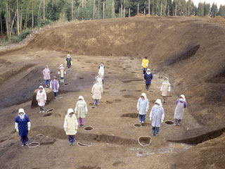 柱の土の窪みの横に16名の作業員の方々が並び、大きさを比較している写真