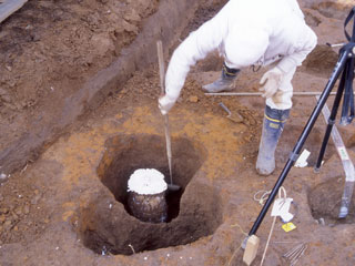 土の中に断面が白い柱の様なものがあり、1名の作業員が柱と側面の土との間に長い金槌のような道具を入れ、見つめている写真