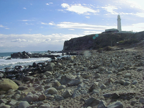 遠くの高台に白い灯台があり、手前には石ころがたくさん転がっている海岸の写真