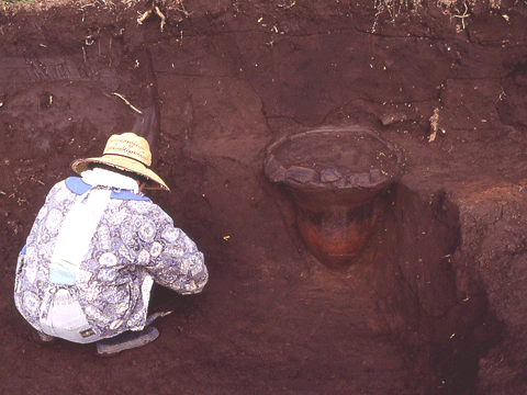 鉢のような大きな土器の横で座って作業する麦わら帽子をかぶった人物の写真